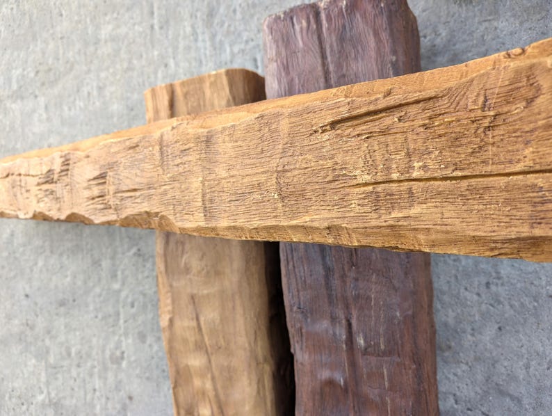 May include: Three rectangular wooden beams arranged in a cross pattern. The horizontal beam is light tan with a textured, weathered grain. The two vertical beams are positioned behind it, one in a light tan shade and the other in a dark brown.