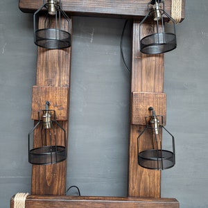 May include: A rustic wooden frame with four black metal cage light fixtures. The frame is made of dark brown wood and has a rope detail at the top. The light fixtures are suspended from the frame and have a black metal cage around the bulb.