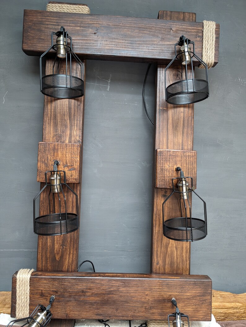 May include: A rustic wooden frame with four black metal cage light fixtures. The frame is made of dark brown wood and has a rope detail at the top. The light fixtures are suspended from the frame and have a black metal cage around the bulb.