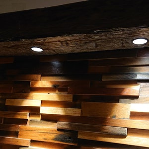 May include: A wooden wall with a rustic, reclaimed wood look. The wall is made up of various sized rectangular pieces of wood, arranged in a staggered pattern. Three recessed spotlights illuminate the wall, casting shadows on the wood.