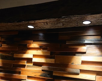 Op de afbeelding: Een houten muur met een rustieke, gerecyclede houten uitstraling. De muur is gemaakt van rechthoekige stukken hout in verschillende maten, die in een verspringend patroon zijn gerangschikt. Drie inbouwspots verlichten de muur en werpen schaduwen op het hout.