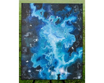 May include: An abstract painting of a nebula in shades of blue and white against a black background. The artwork features swirling cloud-like formations and scattered white stars, creating a cosmic scene. The painting is displayed on a black easel.