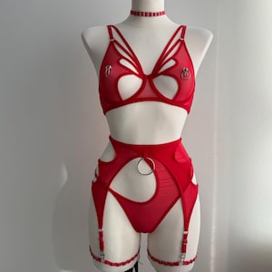 Puede incluir: Conjunto de lencería rojo exhibido en un maniquí. El conjunto incluye un sujetador con diseño de mariposa, una liga a juego, un tanga y un collarín. El sujetador y la liga tienen detalles recortados y herrajes plateados.