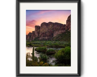 May include: A framed landscape photograph featuring a river flowing past green desert vegetation and a tall saguaro cactus, set against a backdrop of rugged brown mountains under a vibrant sunset sky with hues of orange, pink, and purple.