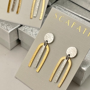 May include: A pair of gold and silver earrings with a geometric design. The earrings are made of two curved pieces of metal, one gold and one silver, that are connected by a smaller piece of metal. The earrings are hanging from a small silver disc.