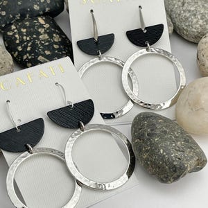 May include: A pair of silver hoop earrings with black leather accents. The hoops are hammered and have a textured finish. The earrings are displayed on a white card with the word "SCAFATI" printed on it.