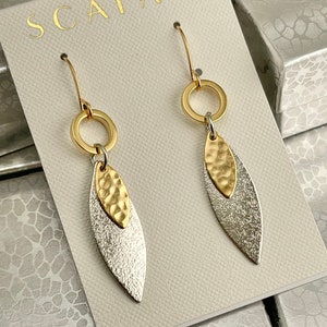 May include: Gold and silver dangle earrings with a hammered leaf design and a circle charm. The earrings are displayed on a white card with the brand name "SCAFATI" printed on it.
