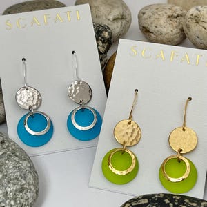 Op de afbeelding: Twee paar oorbellen met een gehamerde metalen schijf en een metalen ring. De oorbellen aan de linkerkant hebben een turquoise blauwe ring en de oorbellen aan de rechterkant hebben een limoengroene ring. De oorbellen worden gepresenteerd op witte kaartjes met het woord "SCAFATI" erop gedrukt.