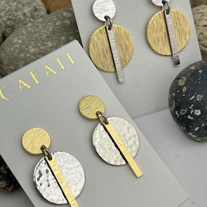 Pode incluir: Brincos pendurados geométricos com esquema de cores dourado e prateado. Cada brinco apresenta um pequeno topo circular, uma grande base circular e uma barra vertical. Os brincos são exibidos num cartão branco com a palavra "CAFATI".