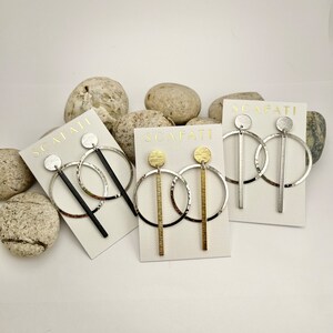 May include: Three pairs of modern dangle earrings on white cards. Each pair features a circular hoop with a vertical bar and a small disc. The earrings are silver, black, and gold-toned. The cards are printed with the word "SCAFATI".