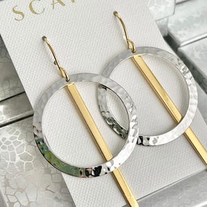 May include: A pair of silver and gold hoop earrings with a hammered texture. The earrings feature a thick gold bar that runs through the center of the hoop.