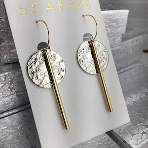 May include: A pair of gold hoop earrings with a silver textured disc and a gold bar hanging from each hoop. The earrings are displayed on a white card with the word "SCAFATI" printed in gold letters.