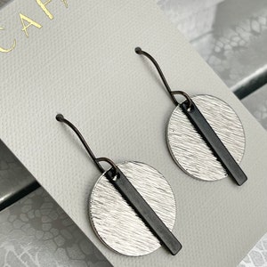 May include: A pair of silver and black dangle earrings with a textured circular design. The earrings are on a white card with the word "CAFATTI" printed in gold.