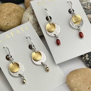 May include: Silver and gold-tone earrings with a circular design. Each earring features a silver disc, a smaller gold disc, and a dangling red or gold bead. The earrings are displayed on white cards with the word "SCAFATI" in gold.