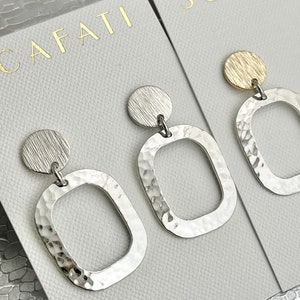 May include: A pair of silver earrings with a hammered texture. Each earring features a round disc at the top and a square-shaped dangle with a rounded edge. The earrings are displayed on a white card with the word "CAFATI" printed in gold.