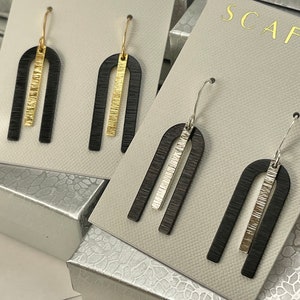 May include: A pair of black arch-shaped earrings with a gold or silver stripe down the centre. The earrings are displayed on a white card with the word "SCAFATI" printed on it.