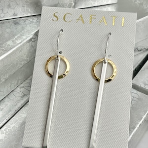 May include: A pair of silver earrings with gold accents. The earrings feature a thin silver bar with a gold circle at the top. The earrings are displayed on a white card with the word "SCAFATI" printed in gold.