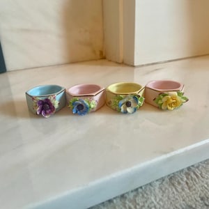 May include: Four pastel-coloured ceramic napkin rings. Each ring is decorated with a floral design in shades of purple, blue, and yellow. The rings are arranged in a row on a white surface. The rings are in light blue, pink, yellow, and pink.