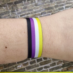 May include: A yellow, white, purple and black ribbon bracelet. The bracelet is tied around a person's wrist.