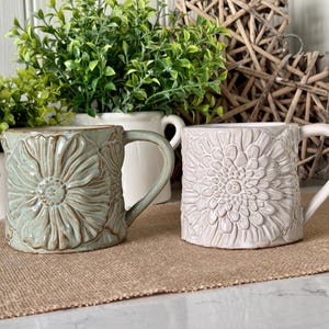 Könnte beinhalten: Zwei Keramikbecher mit floralen Designs. Einer ist hellgrün mit einem Sonnenblumenmuster, der andere ist cremefarben mit einem ähnlichen Blumenmuster. Beide Becher haben Henkel und stehen auf einer braunen Oberfläche.