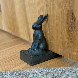 May include: A black cast iron doorstop shaped like a sitting rabbit. The rabbit figure is positioned on a rectangular base and is angled to hold a door open. The surface of the metal has a textured, matte finish, showing the detailed form of the ears.