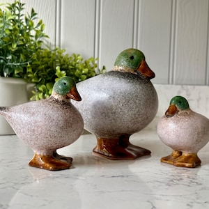Op de afbeelding: Drie keramische eendenbeeldjes met gespikkelde lichamen, groene koppen en bruine snavels. De eenden variëren in grootte, de grootste in het midden. De beeldjes hebben bruine poten en worden tentoongesteld op een wit oppervlak.