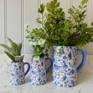 May include: Three ceramic jugs of varying sizes, each featuring a blue and white pattern of rabbits, ferns, and flowers. The jugs have solid blue handles and are filled with green foliage and small white flowers.