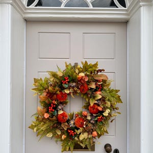 May include: A circular autumn wreath featuring a mix of green and brown foliage, small orange pumpkins, and clusters of bright red berries. The wreath is mounted on a light gray door with a white arched frame and a semi-circular window above.