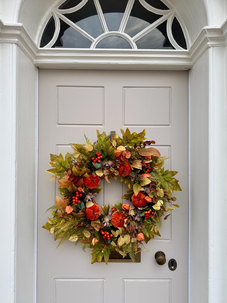 May include: A circular autumn wreath featuring a mix of green and brown foliage, small orange pumpkins, and clusters of bright red berries. The wreath is mounted on a light gray door with a white arched frame and a semi-circular window above.