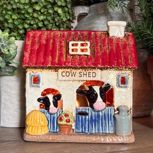 May include: A ceramic cow shed tea light holder with a red roof, cream walls, and the text COW SHED. It features two black and white cows in blue stalls, a yellow hay bale, a potted plant, and a grey milk jug, with a glowing light inside.