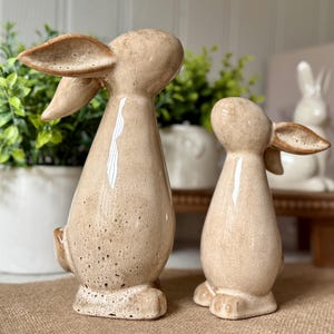 以下が含まれることがあります： 2つのセラミック製のウサギの置物。1つは他方より大きい。どちらも薄い茶色で、斑点のある釉薬がかけられている。大きい方のウサギは耳を立てて直立しており、小さい方のウサギは頭を横に傾けて立っている。