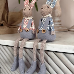 Könnte beinhalten: Zwei Plüschhasen-Figuren mit langen Beinen. Jeder Hase hat einen grauen Körper, eine Latzhose und Filzschuhe. Ein Hase hat rosa geblümte Ohren, der andere blaue. Beide haben einen cremefarbenen Spitzenkragen und Holzknöpfe.