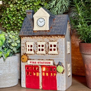 May include: A ceramic fire station building figurine with a blue shingled roof, a clock tower, and red double doors. The facade is light grey with white window frames and the text FIRE STATION in red. A small yellow circle and green leaf are attached.