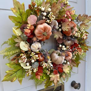 May include: A decorative autumn wreath featuring velvet pumpkins in shades of orange, beige, and brown, accented with green maple leaves, fern fronds, eucalyptus sprigs, and clusters of small white berries, arranged in a circular display.