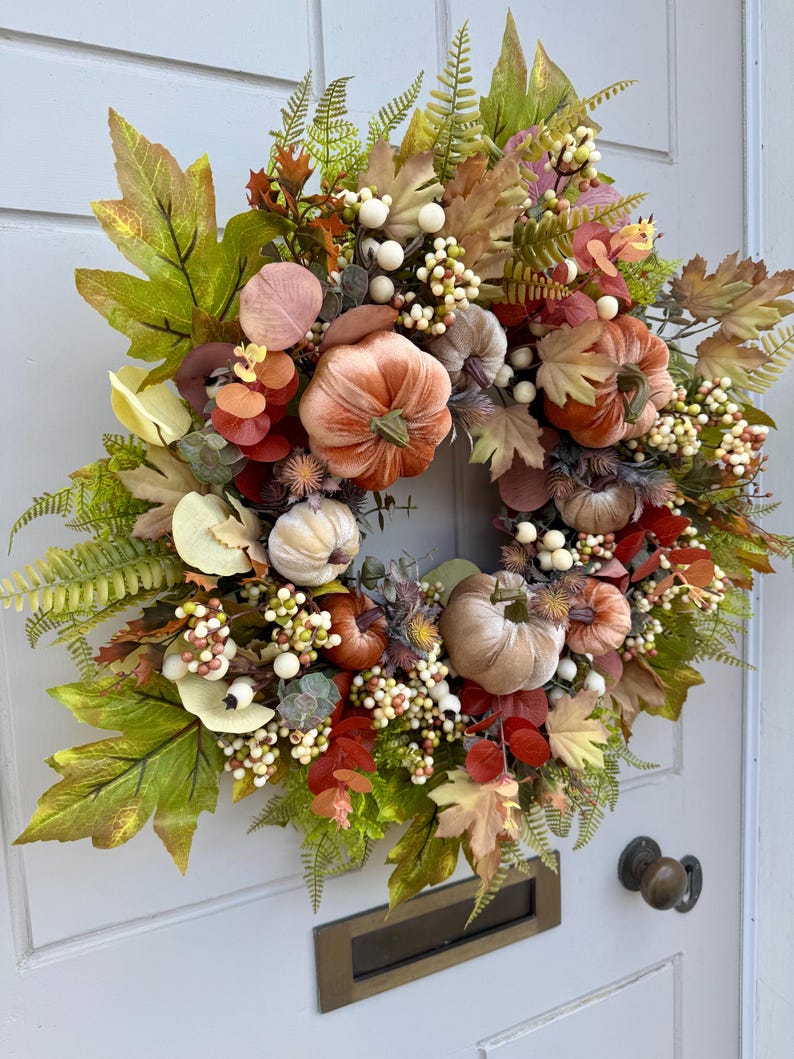 May include: A decorative autumn wreath featuring velvet pumpkins in shades of orange, beige, and brown, accented with green maple leaves, fern fronds, eucalyptus sprigs, and clusters of small white berries, arranged in a circular display.