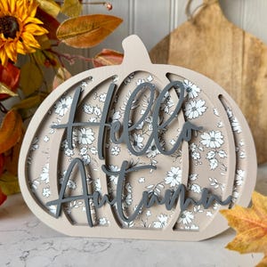 May include: A decorative wooden pumpkin with the words "Hello Autumn" in grey script. The pumpkin features a floral pattern and is surrounded by autumn leaves and sunflowers.