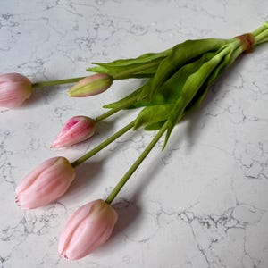 May include: A bundle of artificial tulips with pink petals and green stems and leaves. The tulips are tied together with a brown band. The background is a white marble surface.
