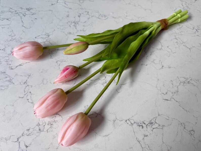 May include: A bundle of artificial tulips with pink petals and green stems and leaves. The tulips are tied together with a brown band. The background is a white marble surface.