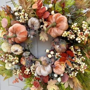May include: A decorative wreath featuring velvet pumpkins in shades of peach and cream, accented with artificial green fern leaves, maple leaves, eucalyptus sprigs, and clusters of small white and tan berries arranged in a circular pattern.