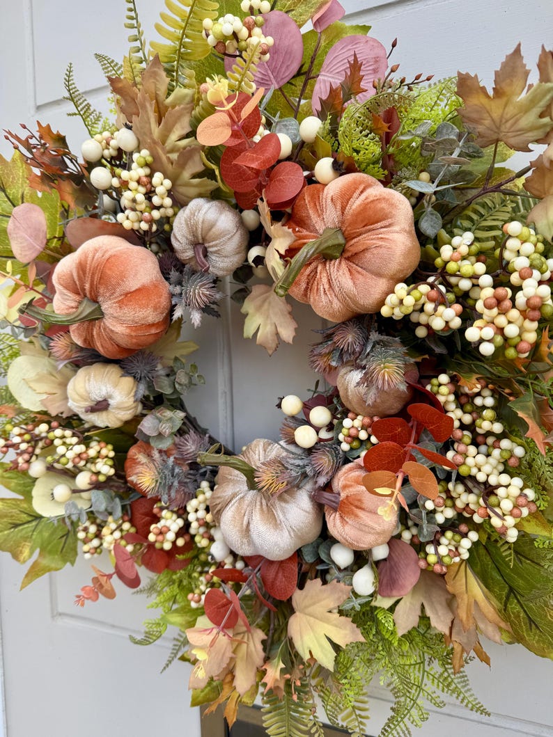 May include: A decorative wreath featuring velvet pumpkins in shades of peach and cream, accented with artificial green fern leaves, maple leaves, eucalyptus sprigs, and clusters of small white and tan berries arranged in a circular pattern.