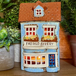 Könnte beinhalten: Ein Teelichthalter aus Keramik in Form eines zweistöckigen Gebäudes mit hellblauer Fassade, braunem Schindeldach und blauer Tür. Die Vorderseite zeigt den Schriftzug THE OLD BAKERY, zwei Fenster mit gelben Blumenkästen und ein leuchtendes Inneres.