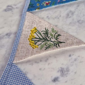May include: Close-up of a handmade bunting banner with triangular fabric flags. The flags feature a variety of patterns, including a floral print, a linen fabric with embroidered yellow flowers and green leaves, and a blue and white gingham check.