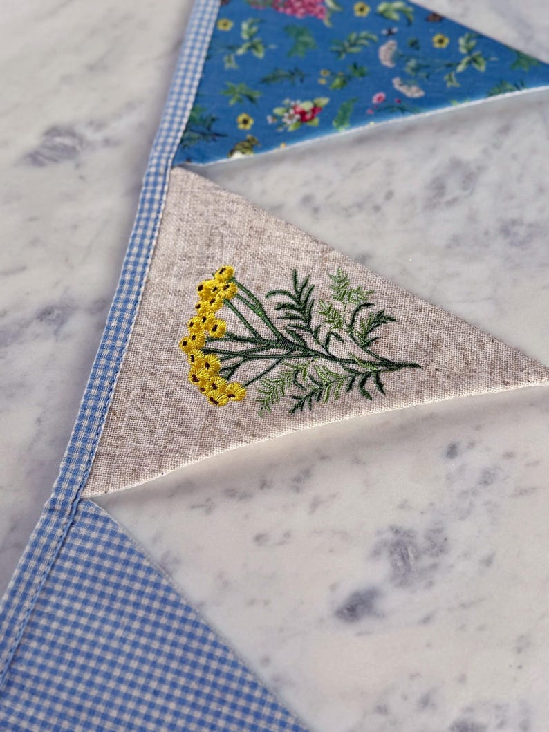 May include: Close-up of a handmade bunting banner with triangular fabric flags. The flags feature a variety of patterns, including a floral print, a linen fabric with embroidered yellow flowers and green leaves, and a blue and white gingham check.