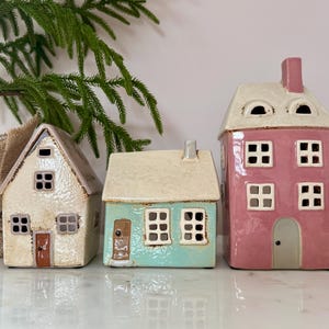 Peut inclure: Trois figurines de maisons en c&eacute;ramique align&eacute;es. Les maisons sont de couleurs diff&eacute;rentes : blanc cass&eacute;, turquoise et rose. Chaque maison a des fen&ecirc;tres carr&eacute;es et une porte. La maison rose est la plus haute et a une chemin&eacute;e. D&eacute;coratif.