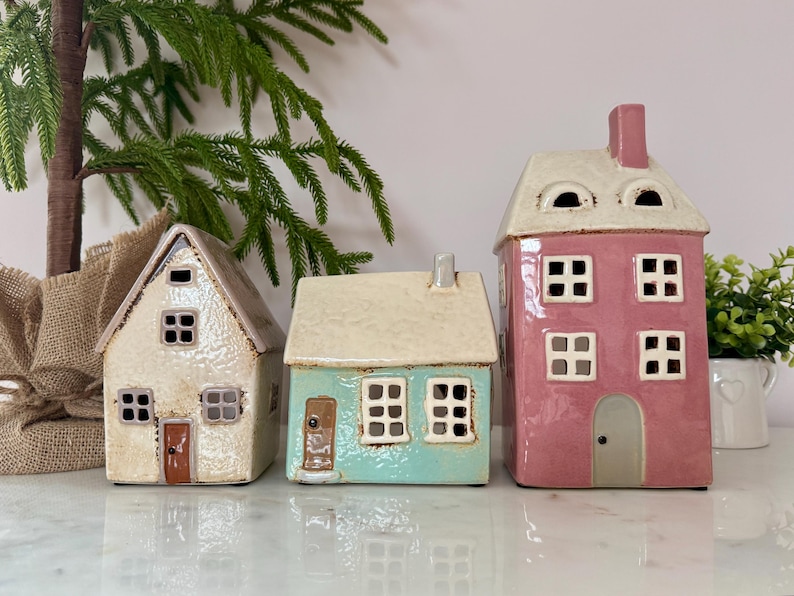Peut inclure: Trois figurines de maisons en c&eacute;ramique align&eacute;es. Les maisons sont de couleurs diff&eacute;rentes : blanc cass&eacute;, turquoise et rose. Chaque maison a des fen&ecirc;tres carr&eacute;es et une porte. La maison rose est la plus haute et a une chemin&eacute;e. D&eacute;coratif.