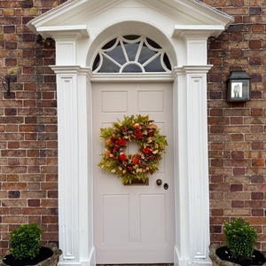 May include: A light beige front door with a six-panel design, featuring a decorative autumn wreath made of green, orange, and red leaves and small pumpkins. The door is framed by white architectural molding and a semi-circular fanlight window above.