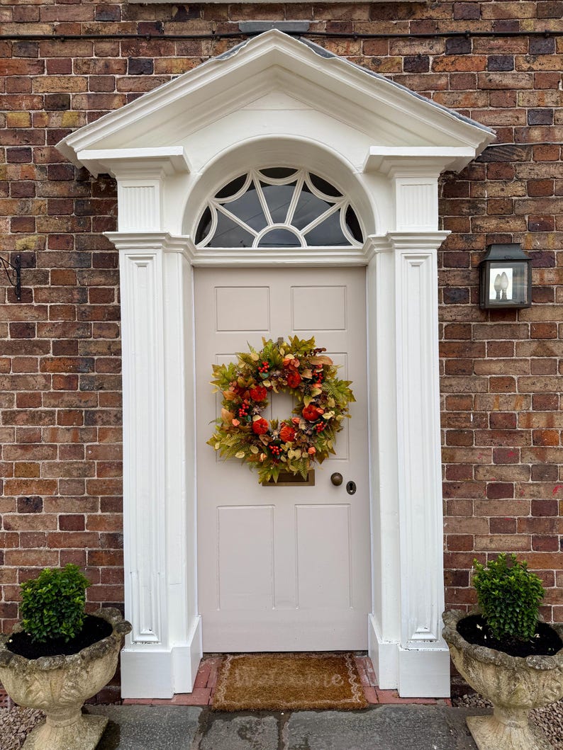 May include: A light beige front door with a six-panel design, featuring a decorative autumn wreath made of green, orange, and red leaves and small pumpkins. The door is framed by white architectural molding and a semi-circular fanlight window above.
