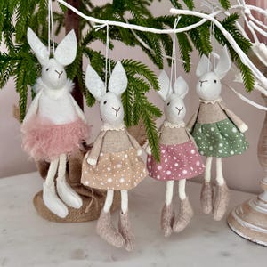 Könnte beinhalten: Vier handgefertigte Hasenornamente mit langen Ohren und hängenden Beinen. Die Hasen tragen Kleider in Rosa, Beige und Grün mit weißen Punkten. Ein Hase hat einen flauschigen rosa Rock. Sie hängen an weißen Schnüren vor einem Hintergrund aus Grün.