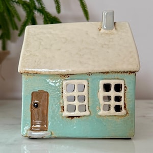 Peut inclure: Une maison en c&eacute;ramique turquoise avec un toit cr&egrave;me et une petite chemin&eacute;e. La maison pr&eacute;sente une porte marron et deux fen&ecirc;tres avec des ouvertures carr&eacute;es. C'est un objet d&eacute;coratif.