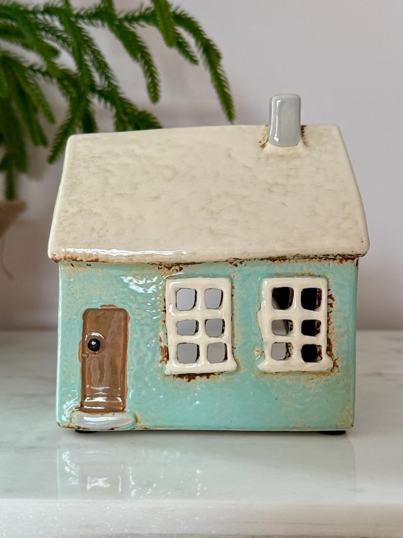 Peut inclure: Une maison en c&eacute;ramique turquoise avec un toit cr&egrave;me et une petite chemin&eacute;e. La maison pr&eacute;sente une porte marron et deux fen&ecirc;tres avec des ouvertures carr&eacute;es. C'est un objet d&eacute;coratif.